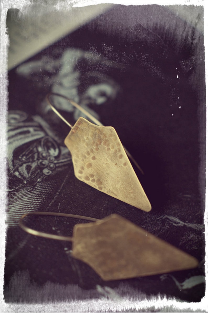 May include: A pair of gold-toned earrings with a hammered diamond shape. The earrings are hanging from a dark background.