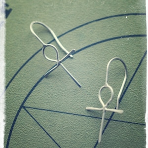 Peut inclure: Une paire de boucles d'oreilles Ankh en argent sur un fond vert avec des lignes noires.