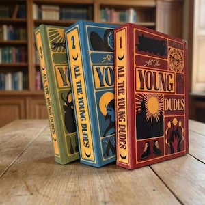 Op de afbeelding: Drie gebonden boeken getiteld All The Young Dudes, genummerd van één tot drie. Het rode boek toont een trein en zon, het blauwe een maan en tent, en het groene een hert-silhouet. Op elke rug staat de titel van het boek vermeld.