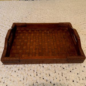 Puede incluir: Una bandeja de servicio rectangular de madera marrón con un patrón de tejido de cesta en la base. La bandeja incluye asas laterales integradas y detalles decorativos envueltos en cada una de las cuatro esquinas, lo que le da un aspecto estructurado.