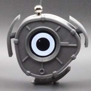 Könnte beinhalten: Ein graues, 3D-gedrucktes, dreieckiges elektronisches Gerät mit einem zentralen kreisförmigen Bildschirm, der eine schwarze Pupille auf weißem Hintergrund zeigt. Das Objekt hat einen silbernen Knopf oben und drei geschwungene, flügelartige Vorsprünge.