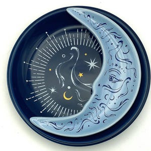 May include: A round, dark blue ceramic dish featuring a light blue crescent moon overlay. The dish interior displays a white sunburst pattern, small yellow stars, and a small yellow crescent moon against a deep navy background.