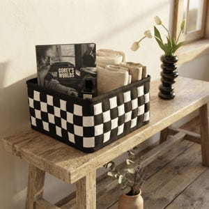May include: A square storage basket featuring a black and white woven checkerboard pattern. Inside the basket are folded beige textiles, a book titled Gorey's Worlds, and a small dark bottle. The basket is constructed from thick fabric strips.