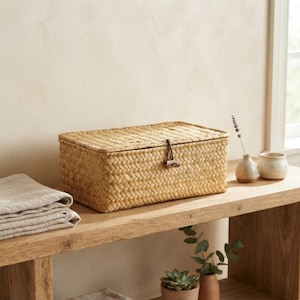 May include: A rectangular woven seagrass storage box with a matching lid. The lid is secured by a dark brown elastic cord looped over a small, dark wooden toggle button centered on the front of the box. The weave is a consistent light tan color.