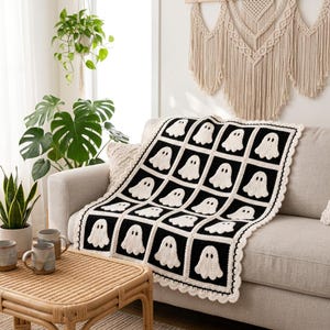 May include: A crochet blanket featuring a repeating grid pattern of white ghost motifs on black square backgrounds. The blanket is finished with a scalloped white border and is draped over a neutral-colored sofa.