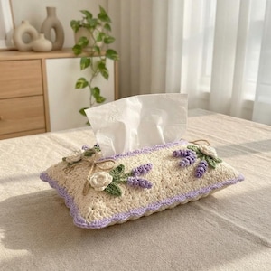 Könnte beinhalten: Ein rechteckiger Taschentuchbox-Überzug aus cremefarbenem Häkelgarn mit einem lavendelfarbenen Rand. Der Überzug ist mit dreidimensionalen gehäkelten weißen Blumen, grünen Blättern und violetten Lavendelzweigen sowie einer kleinen Juteschleife verziert.