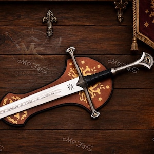 May include: A silver-toned metal sword with a black wrapped handle and a star emblem on the blade, featuring engraved runic text. The sword is mounted on a brown wooden plaque with gold-coloured floral patterns and a curved, shield-like shape.