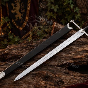 May include: A silver-coloured metal sword with a black leather-wrapped grip and a cross-shaped guard. The blade features engraved runic text along its length. Beside the sword lies a black scabbard with silver-coloured metal fittings at both ends.