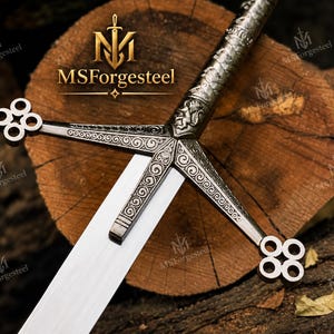 May include: A silver-toned metal sword featuring an intricate crossguard with circular quatrefoil terminals and engraved scrollwork patterns. The hilt displays a relief of a lion, and the blade is straight with a polished finish. The text MSForgesteel is visible.