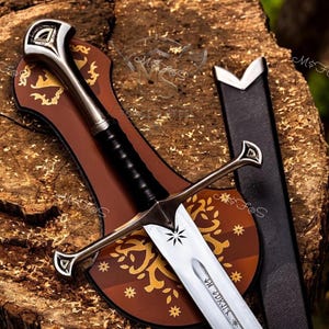 May include: A silver-toned metal sword with a black grip, a cross-shaped guard, and a blade featuring a star engraving and runic text. The sword is accompanied by a brown shield-shaped plaque with gold floral patterns and a black leather scabbard.