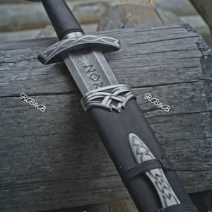 May include: A sword with a dark wooden handle and a silver pommel featuring intricate engravings. The blade is partially visible, displaying the letters NOR, and is housed in a dark wooden scabbard adorned with silver metal fittings and knotwork.