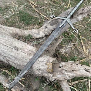 May include: A long sword with a distinctive Damascus steel blade featuring a wavy, layered pattern. The metal crossguard and pommel are intricately engraved with floral motifs, and the grip is wrapped in a matching silver-toned metal finish.