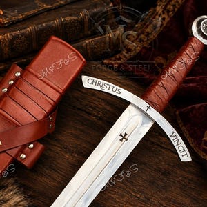 May include: A steel sword with a brown leather-wrapped handle and a matching brown leather scabbard. The crossguard is engraved with the text CHRISTUS VINCIT and a small cross. The scabbard features two gold-coloured cross emblems.