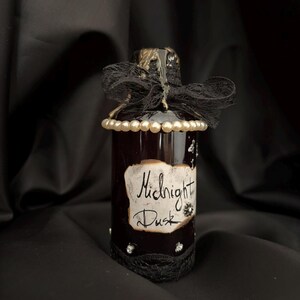 Può includere: Una bottiglia in vetro marrone scuro con un fiocco in pizzo nero sul collo, una fila di perle color crema e un'etichetta di carta con bordi bruciati che riporta la scritta Midnight Dusk in corsivo nero. Sono presenti piccoli dettagli argentati.