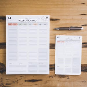 Peut inclure: Un bloc-notes hebdomadaire A4 et un A5 avec du papier blanc, des lignes de grille grises et des en-têtes de couleur pastel pour les jours de la semaine. Chaque planificateur comporte des sections pour les tâches, notes et objectifs. Un stylo bille gris foncé est placé entre les deux.