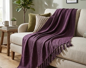 Puede incluir: Una manta tejida de color morado intenso con una superficie texturizada y bordes con flecos anudados, colocada sobre un sofá de color beige claro. La manta presenta un patrón de tejido denso y uniforme que cuelga por el frente.
