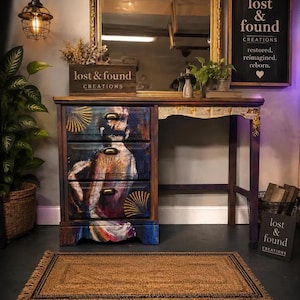 May include: A wooden desk featuring three drawers with brass pulls, decorated with a colorful artistic print of a figure. The desk includes a gold-framed mirror, a wooden sign reading lost & found CREATIONS, and a rectangular woven rug.