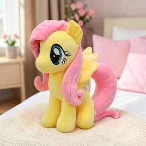 May include: A plush toy of a yellow pony with a long pink mane and tail. The pony has large teal eyes with long eyelashes, small yellow wings, and a cutie mark featuring three pink butterflies on its flank. The toy is made of soft fabric.