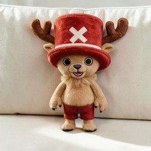 May include: A plush toy of a tan reindeer character with large brown antlers, wearing a tall red top hat featuring a white cross symbol on the front. The figure has a light tan furry chest, a blue nose, and is dressed in red shorts with black feet.
