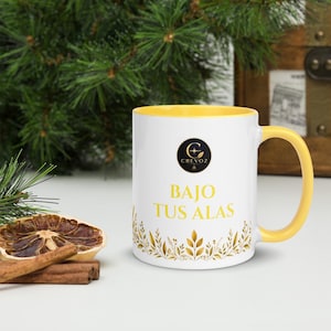 May include: A white ceramic mug with a yellow interior and handle. The front features a black circular logo with the text CREVOZ, followed by the yellow text BAJO TUS ALAS, and a decorative border of golden leaves at the base of the mug.