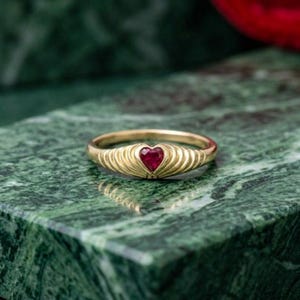 Puede incluir: Un anillo de oro con una piedra preciosa roja en forma de corazón en el centro. La banda está diseñada con una serie de crestas verticales en relieve que se abren en abanico desde el centro, creando una apariencia texturizada de concha.