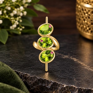 Puede incluir: Un anillo de oro con tres piedras preciosas de peridoto de corte ovalado alineadas verticalmente, cada una acentuada con una pequeña piedra preciosa redonda de color rosa. El diseño incluye una barra de oro vertical central que se extiende más allá de las piedras.