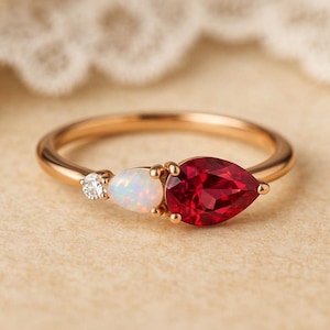 May include: A gold ring featuring a pear-shaped red gemstone, a smaller oval-shaped iridescent opal, and a tiny round clear diamond-like stone. The band is polished gold, and the stones are set in a horizontal arrangement along the ring band.