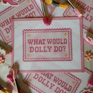 May include: A rectangular needlepoint canvas featuring a pink gingham border and the text "WHAT WOULD DOLLY DO?" in pink cross-stitch lettering. The design includes small yellow star accents within a decorative pink frame, surrounded by embroidery floss.