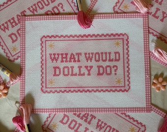 May include: A rectangular needlepoint canvas featuring a pink gingham border and the text "WHAT WOULD DOLLY DO?" in pink cross-stitch lettering. The design includes small yellow star accents within a decorative pink frame, surrounded by embroidery floss.