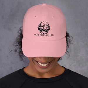 May include: A light pink baseball cap featuring a black graphic of a curly-haired dog's head centered on the front panel. Below the dog illustration, the text reads LEAD AND LOOM CO. in a simple, bold, black sans-serif font.