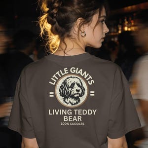 Könnte beinhalten: Ein dunkelgraues T-Shirt mit einer kreisförmigen Grafik auf der Rückseite. Das Design zeigt eine schwarz-weiße Illustration eines Hundekopfes, umgeben von dem Text "LITTLE GIANTS", "LIVING TEDDY BEAR" und "100% CUDDLES" in cremefarbener Schrift.