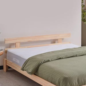 Könnte beinhalten: Ein helles Holzbettgestell mit einem Kopfteil aus horizontalen Latten. Das Bett ist mit einer weißen Matratze und einem strukturierten olivgrünen Bettbezug ausgestattet. Ein kleiner runder Beistelltisch aus Holz steht neben dem Bettgestell.