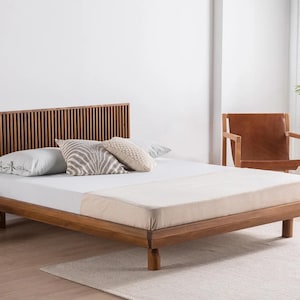 Op de afbeelding: Een laag houten bedframe met een hoofdeinde van verticale latten. Het bed is opgemaakt met een wit hoeslaken en drie sierkussens, waaronder één met een grijze print en twee met een gestructureerde stof in neutrale tinten.