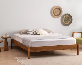 May include: A wooden bed frame with a white mattress, topped with two beige pillows, a striped rectangular pillow, and a round white pom-pom pillow. A light gray throw blanket is draped across the foot of the bed, which is set in a room.