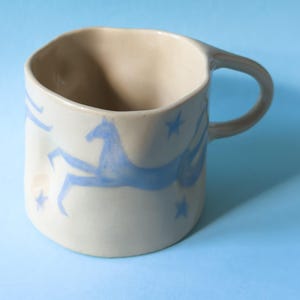 May include: A beige ceramic mug with an irregular, hand-formed shape and a single handle. The exterior features a hand-painted design of a blue horse in motion surrounded by small blue stars, creating a whimsical celestial scene on the surface.