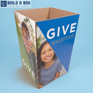 May include: A square cardboard donation bin featuring a blue panel with the white text GIVE and Thank you for HELPING CHILDREN AND FAMILIES IN OUR COMMUNITY. The side panels display photographic portraits of a young boy and a young girl smiling.