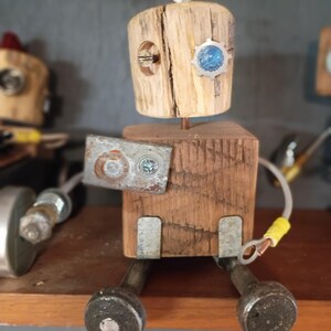 以下が含まれることがあります： 木製のブロックの頭と胴体で作られた小さなロボットのフィギュア。青い円形の目、胸の金属プレート、金属製のボルトの足が特徴です。頭の上には銀色のリングがあり、黄色いコネクタが付いたワイヤーの腕が取り付けられています。