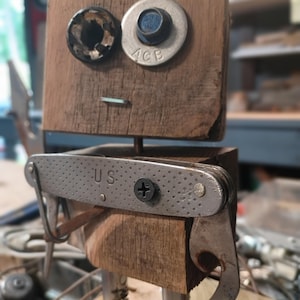 以下が含まれることがあります： 2つの木製ブロックで作られたロボットのフィギュア。頭部には暗い金属製のワッシャーと銀色のボルトが目として配置され、小さな金属製のピンが口として付いています。胴体は木製のブロックで、USと刻印された銀色のマルチツールと黒いネジで装飾されています。
