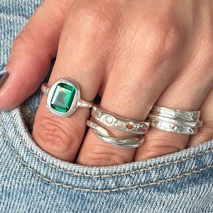 Hand tucked into a light denim pocket wearing five fine silver rings: a large green emerald-cut stone in a rustic bezel, a wide molten-textured band set with tiny clear and amber stones, a twisted wrap ring and two hammered stacking bands.