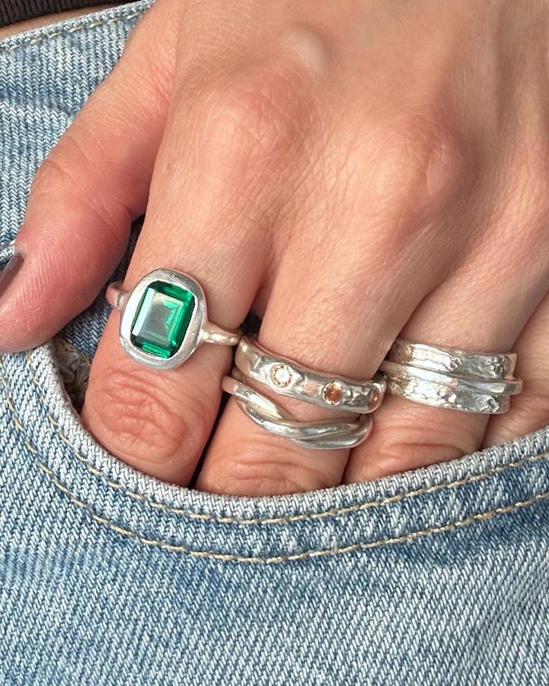 Hand tucked into a light denim pocket wearing five fine silver rings: a large green emerald-cut stone in a rustic bezel, a wide molten-textured band set with tiny clear and amber stones, a twisted wrap ring and two hammered stacking bands.