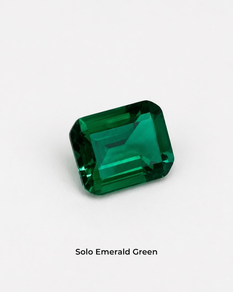 A single deep green emerald-cut stone, 8 x 10 mm, rectangular with cut corners and flat step facets, on a pale grey background. Shown alone at roughly the width of a fingernail. Labelled Solo Emerald Green.