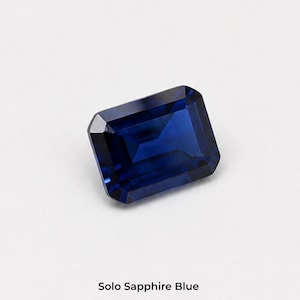 A single deep blue emerald-cut stone, 8 x 10 mm, rectangular with cut corners and flat step facets, on a pale grey background. Shown alone at roughly the width of a fingernail. Labelled Solo Sapphire Blue.