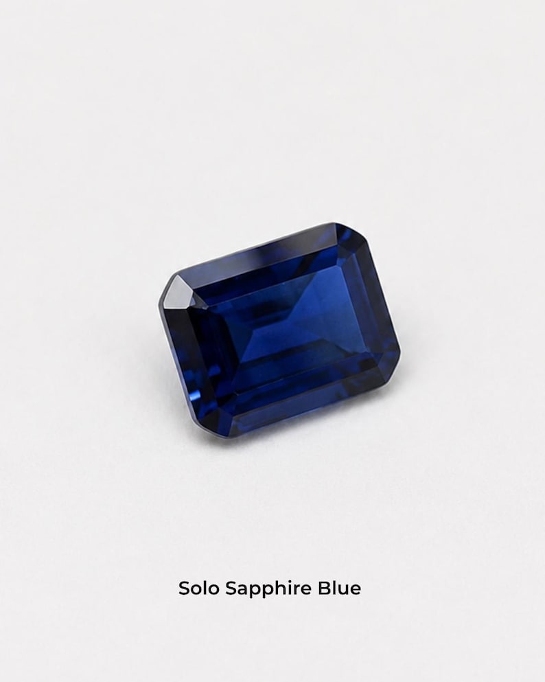 A single deep blue emerald-cut stone, 8 x 10 mm, rectangular with cut corners and flat step facets, on a pale grey background. Shown alone at roughly the width of a fingernail. Labelled Solo Sapphire Blue.