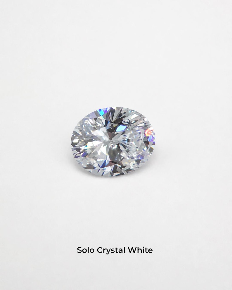 A single clear colourless oval brilliant-cut stone, 9 x 11 mm, on a pale grey background, its facets throwing small flashes of blue and orange. Shown alone at roughly the width of a fingernail. Labelled Solo Crystal White.