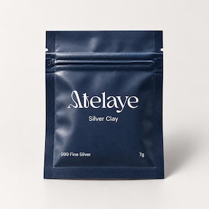 Pode incluir: Uma bolsa retangular selada de cor azul escuro contendo argila de prata. A frente apresenta o texto branco 'Atelaye', 'Silver Clay', '999 Fine Silver' e '7g'. A embalagem tem um fecho hermético horizontal e uma borda texturizada.