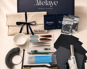 May include: A metal clay jewelry making kit featuring a dark blue box labeled Atelaye, a beige fabric pouch with a blue ribbon, silver clay packets, a white ceramic crucible, metal tweezers, sculpting tools, a ring sizer, brushes, and a butane torch.