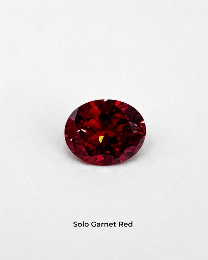 A single garnet red oval brilliant-cut stone, 9 x 11 mm, on a pale grey background. Shown alone at roughly the width of a fingernail. Labelled Solo Garnet Red.