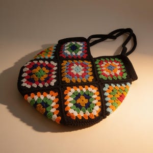 Puede incluir: Un bolso de ganchillo construido a partir de múltiples motivos cuadrados con un borde de hilo negro. Cada cuadrado muestra un patrón vibrante y multicolor en tonos rojo, azul, verde, amarillo, naranja y blanco. El bolso incluye dos asas negras.