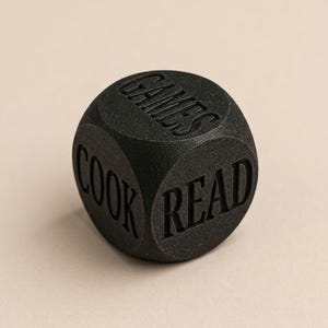 Könnte beinhalten: Ein schwarzer sechsseitiger Würfel mit strukturierter, matter Oberfläche. Jede Seite zeigt ein eingeprägtes Wort in Großbuchstaben. Die sichtbaren Seiten zeigen die Wörter GAMES, COOK und READ, die deutlich in die Oberfläche des Würfels eingraviert sind.