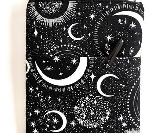 Puede incluir: Bolsa negra con cremallera con un diseño celestial con lunas, estrellas y soles blancos. Un cuaderno floral asoma por la parte superior. Un bolígrafo está en el bolsillo delantero. La bolsa está diseñada para guardar un cuaderno o una tableta.